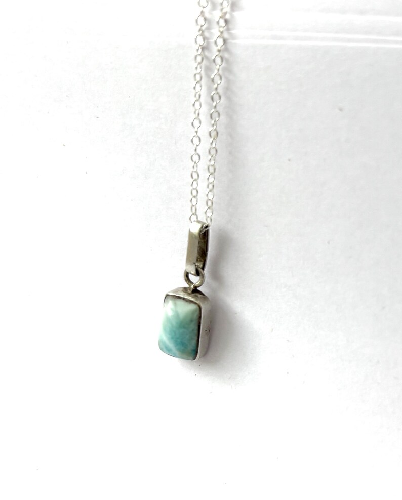 Op de afbeelding: Een zilveren ketting met een rechthoekige hanger met een lichtblauwe en witte steen. De hanger hangt aan een fijne zilveren ketting. De steen heeft een gemarmerd uiterlijk en de zetting is van gepolijst zilver.