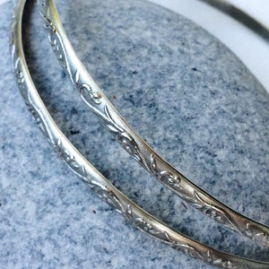 May include: Two silver bangles with a floral design. The bangles are stacked on top of each other.