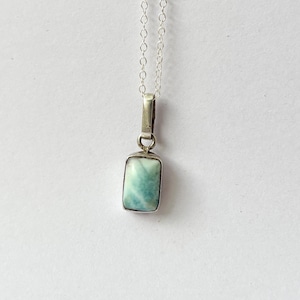 May include: A rectangular pendant necklace with a light blue gemstone set in a silver-colored metal frame. The pendant hangs from a delicate silver chain. The gemstone has a marbled appearance with shades of blue and white.