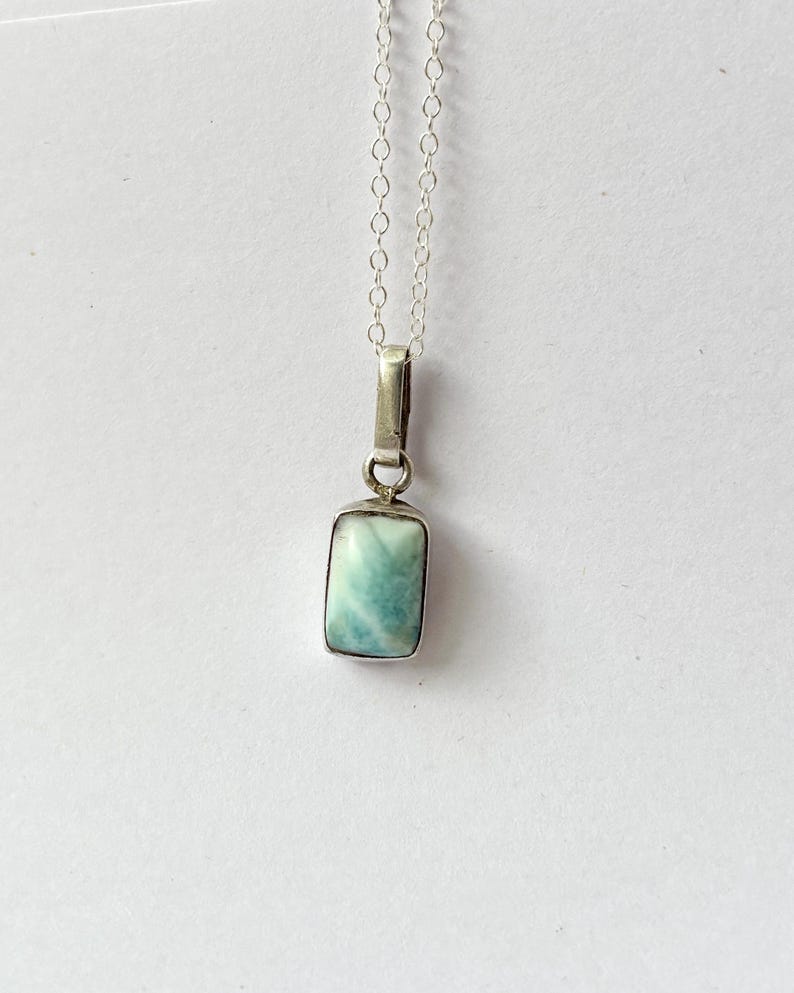 Op de afbeelding: Een rechthoekige hangerketting met een lichtblauwe edelsteen gezet in een zilverkleurig metalen frame. De hanger hangt aan een fijne zilveren ketting. De edelsteen heeft een gemarmerd uiterlijk met tinten blauw en wit.
