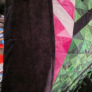 Puede incluir: Primer plano de un edred&oacute;n con borde marr&oacute;n oscuro. El edred&oacute;n presenta un dise&ntilde;o geom&eacute;trico con tri&aacute;ngulos rosas, blancos y verdes. Los tri&aacute;ngulos rosas tienen un estampado floral. El edred&oacute;n est&aacute; doblado.