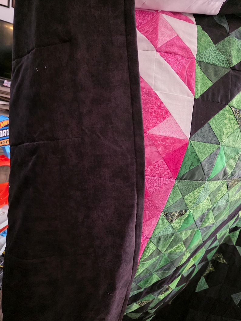 Puede incluir: Primer plano de un edred&oacute;n con borde marr&oacute;n oscuro. El edred&oacute;n presenta un dise&ntilde;o geom&eacute;trico con tri&aacute;ngulos rosas, blancos y verdes. Los tri&aacute;ngulos rosas tienen un estampado floral. El edred&oacute;n est&aacute; doblado.