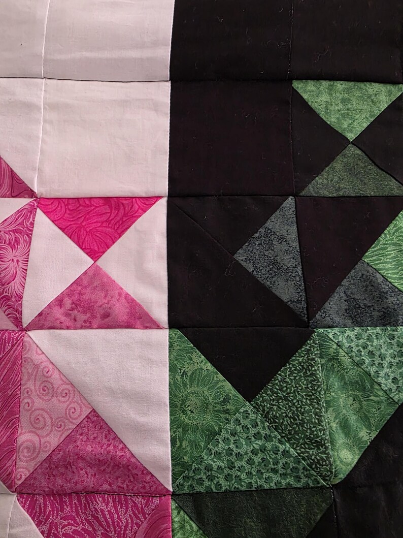 Puede incluir: Primer plano de un edred&oacute;n de patchwork con un dise&ntilde;o geom&eacute;trico. El edred&oacute;n presenta cuadrados de telas blancas, negras y rosas. La tela rosa tiene un patr&oacute;n de remolino. Tambi&eacute;n hay tri&aacute;ngulos verdes con patrones florales y de hojas.