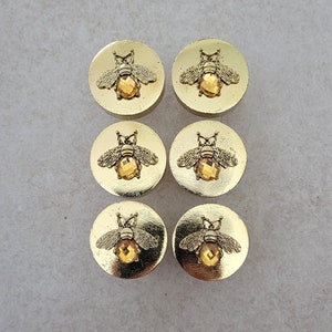 May include: Six gold-toned round buttons with a bee design and a clear rhinestone in the centre.