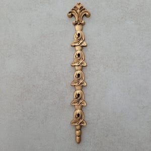 May include: A gold-coloured, vertical decorative element featuring a fleur-de-lis at the top and a series of rose-like ornaments. The design is symmetrical and detailed, with a textured surface. The bottom has small, rounded shapes.