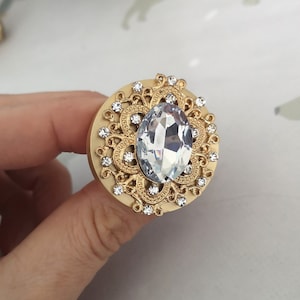 Op de afbeelding: Een gouden decoratieve knop met een groot, helder, ovaalvormig kristal in het midden. Het kristal wordt omringd door een gedetailleerd, versierd ontwerp van kleinere kristallen en gouden filigraan.