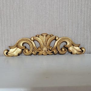 May include: A gold-coloured decorative trim with a scroll design. The trim has a central motif with a stylised leaf shape and two smaller scrolls on either side.