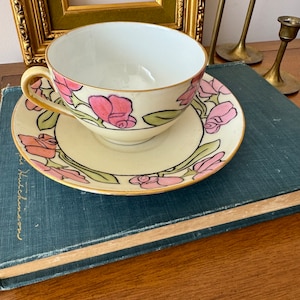 May include: A vintage teacup and saucer set with a floral design. The cup and saucer are white with a gold trim and feature pink roses with green leaves.