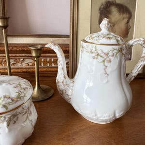 May include: White porcelain coffee pot with a lid, featuring delicate floral patterns in pink and green, accented with gold trim. The pot has a curved spout and handle, and sits on a wooden surface. A matching sugar bowl is also visible.