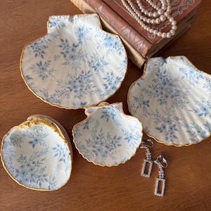 May include: Three white ceramic seashell-shaped dishes with a blue floral pattern and gold trim. The dishes are arranged on a wooden surface with a pair of silver earrings.
