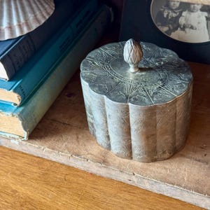 May include: A silver, round, decorative box with an ornate design and a pinecone-shaped knob on the lid.