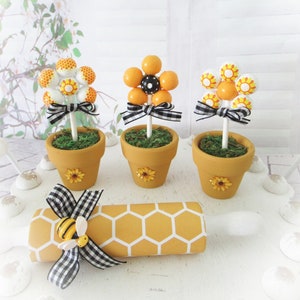 May include: Three small potted plants with yellow and orange flowers made from round balls. The pots are decorated with sunflowers. A yellow and white rolling pin with a honeycomb pattern and a black and white checked bow with a bee charm is in the foreground.