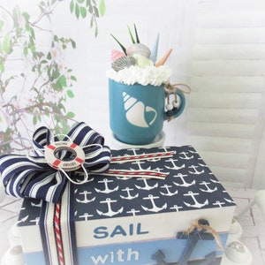 Op de afbeelding: Een blauwe keramische mok met een wit schelpmotief en een witte rand. De mok is gevuld met witte slagroom en bedekt met schelpen, zeesterren en een klein groen plantje. De mok staat op een blauwe en witte doos met een wit ankerpatroon en de tekst "SAIL with".
