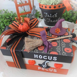 May include: A black and orange gift box with a black ribbon and a pumpkin on top. The box is decorated with a witch's hat, a broom, and the words "Hocus Pocus".