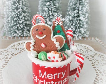 Puede incluir: Una taza roja con el texto blanco Merry Christmas, llena de dulces navideños decorativos de imitación que incluyen un hombre de jengibre, un árbol verde, un bastón de caramelo y pequeños dulces redondos, con una cinta a rayas rojas y blancas en el asa.