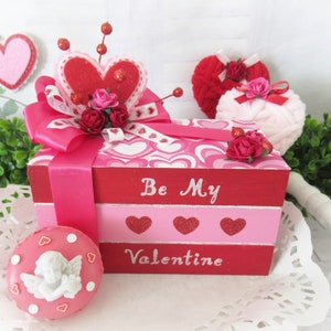 Op de afbeelding: Een roze en rode houten doos met een hartvormig ontwerp en de woorden "Be My Valentine" op de voorkant. De doos is versierd met een roze lint, een rood vilt hart en roze bloemen. Er is een klein roze en wit engelenbeeldje op een roze en witte stippenbasis.