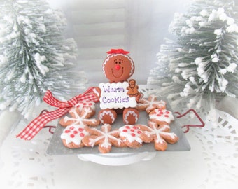 May include: A gingerbread person holding a sign that says "Warm Cookies" is surrounded by gingerbread snowflake cookies on a silver tray. The tray is on a white cake stand. The gingerbread person is wearing a red bow.