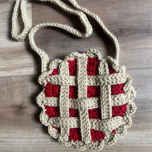 May include: A crocheted pie-shaped bag with a red and beige pattern. The bag has a long beige strap.