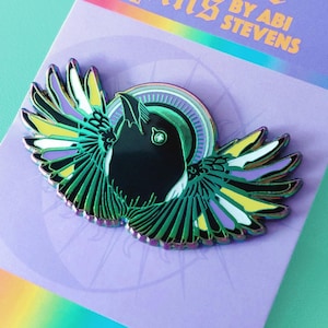 May include: A metal enamel pin featuring a black bird with outstretched wings in shades of green, yellow, and white, mounted on a purple backing card with the text Pride Pins by Abi Stevens and a rainbow gradient stripe at the bottom.
