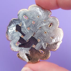 May include: A detailed, flower-shaped enamel pin with a gold outline. The pin features a complex design with black and white patterns, held between two fingers against a purple background. The pin's design includes a repeating pattern.