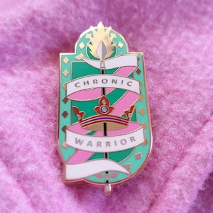 May include: A decorative enamel pin with the text "CHRONIC WARRIOR". The pin has a shield shape with a teal background, pink and white ribbons, a gold crown, and a spear. The pin is set against a pink fabric background.