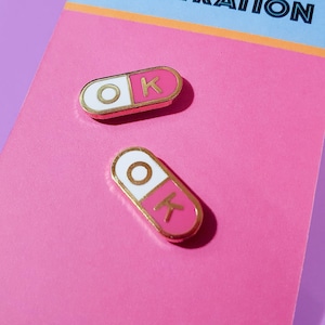 A pair of matching 0.75 inch mini enamel pins in the shape of a pill; half pink, half white, with the letters O and K inside the halves. They are hard-enamel (a smooth surface, not recessed enamel), and have gold plating on the metal.