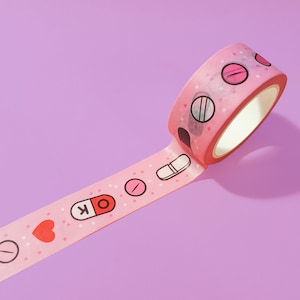 May include: Pink washi tape with a repeating pattern of pills, hearts, and white dots. The tape is rolled up and partially unrolled, showing the pattern.