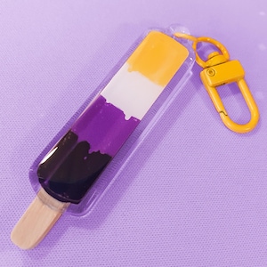 Peut inclure: Un porte-clés en forme de glace à l'eau aux couleurs du drapeau de la fierté non binaire : jaune, blanc, violet et noir. Le porte-clés a un bâton en bois et un fermoir jaune. Le fond est une surface violet clair avec des points blancs.