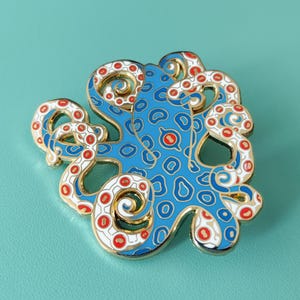 May include: A gold-toned metal enamel pin shaped like an octopus. The octopus features a bright blue body with a darker blue spotted pattern and white tentacles adorned with small red circular accents. The design has a polished gold metal outline.