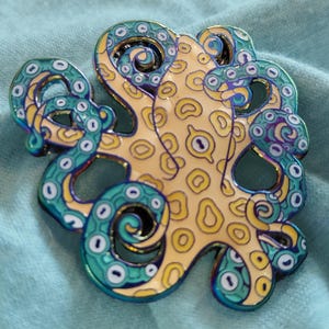 May include: A metal enamel pin shaped like an octopus with a cream-coloured head featuring yellow spots and teal tentacles with white suction cups. The metal edges of the pin have an iridescent, rainbow-coloured finish that reflects light.