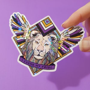 Puede incluir: Una pegatina holográfica con un león blanco con alas. Las alas están formadas por rayas de morado, amarillo y negro. El león está en un marco en forma de diamante con el texto "I found my pride" en la parte inferior.