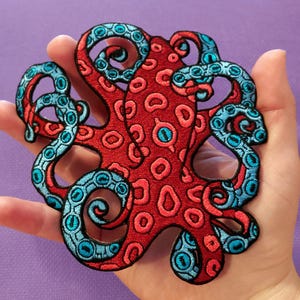 Peut inclure: Un écusson brodé en forme de pieuvre avec un corps rouge orné de motifs circulaires roses et des tentacules bleu vif aux contours sombres. Le design présente des détails de couture visibles sur l'ensemble, mettant en valeur les bras enroulés.
