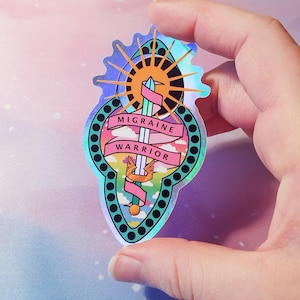 Pu&ograve; includere: Un adesivo olografico con un design arcobaleno che presenta una spada, un sole e nuvole. Il testo "Migraine Warrior" &egrave; scritto su uno stendardo rosa.