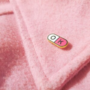 One of the enamel pins photographed on a light pink jacket coller. The pill shaped enamel pin is split in 2, with the wordK OK in the halves; O in the white half and K in the pink half.