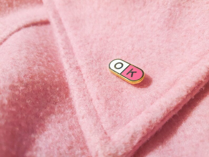 One of the enamel pins photographed on a light pink jacket coller. The pill shaped enamel pin is split in 2, with the wordK OK in the halves; O in the white half and K in the pink half.