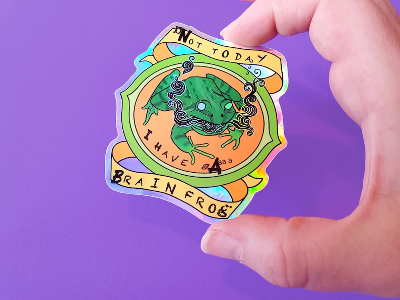 May include: A holographic sticker with a green frog with a blue and black smoke design. The sticker says "Not Today I Have Brain Frog".
