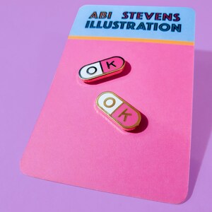 A pair of matching 0.75 inch mini enamel pins in the shape of a pill; half pink, half white, with the letters O and K inside the halves. They are hard-enamel (a smooth surface, not recessed enamel), and have gold plating on the metal.