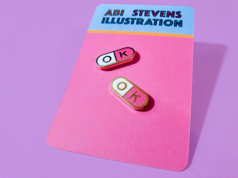 A pair of matching 0.75 inch mini enamel pins in the shape of a pill; half pink, half white, with the letters O and K inside the halves. They are hard-enamel (a smooth surface, not recessed enamel), and have gold plating on the metal.