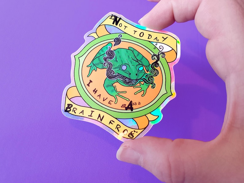 May include: A holographic sticker with a green frog with a blue eye and a black mustache. The frog is surrounded by a yellow, orange, and green circle with the text "I have a brain frog" and "Not today".