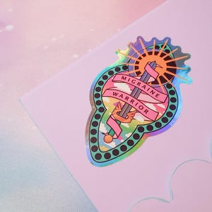 Pu&ograve; includere: Un adesivo olografico con un design rosa e verde. Presenta una spada con ali, un sole e nuvole. Il testo "Migraine Warrior" &egrave; scritto su uno stendardo rosa.