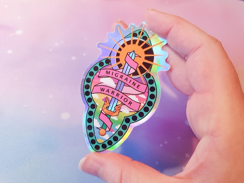 Pu&ograve; includere: Un adesivo olografico con una spada, un sole e nuvole. Il testo "Migraine Warrior" &egrave; scritto su uno stendardo rosa.