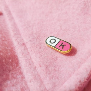 One of the enamel pins photographed on a light pink jacket coller. The pill shaped enamel pin is split in 2, with the wordK OK in the halves; O in the white half and K in the pink half.