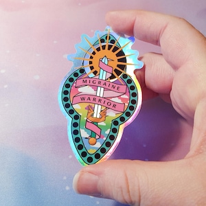 Pu&ograve; includere: Un adesivo olografico con una spada, un sole e nuvole. Il testo "Migraine Warrior" &egrave; su uno stendardo rosa.