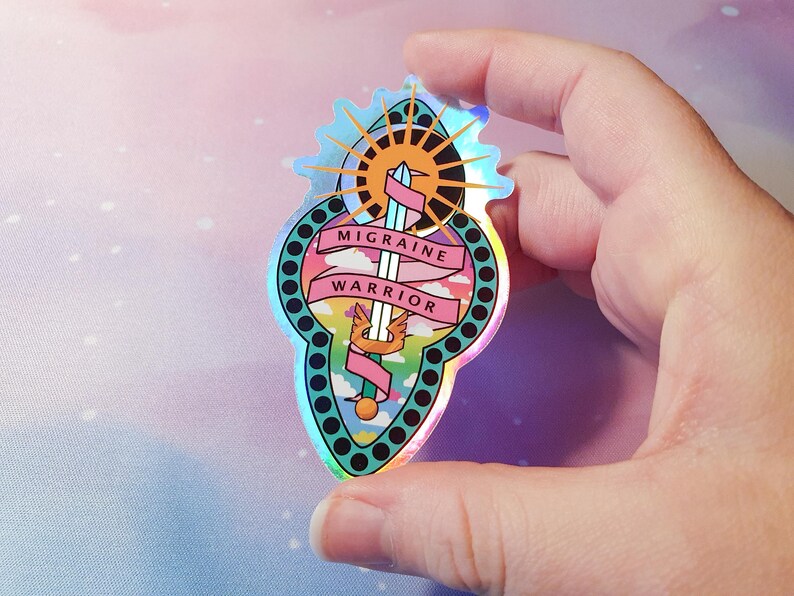 Pu&ograve; includere: Un adesivo olografico con una spada, un sole e nuvole. Il testo "Migraine Warrior" &egrave; su uno stendardo rosa.
