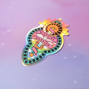 Pu&ograve; includere: Un adesivo olografico con un design colorato di una spada con ali e un sole. Il testo "Migraine Warrior" &egrave; scritto su uno stendardo attorno alla spada.
