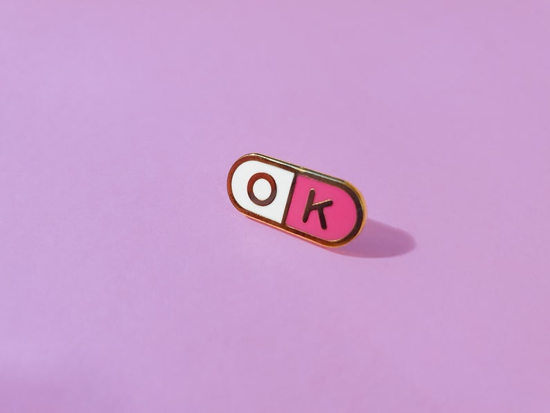 One of a pair of matching 0.75 inch; mini enamel pins in the shape of a pill; half pink, half white, with the letters O and K inside the halves. They are hard-enamel (a smooth surface, not recessed enamel), and have gold plating on the metal.