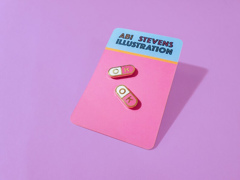 A pair of matching 0.75 inch mini enamel pins in the shape of a pill; half pink, half white, with the letters O and K inside the halves. They are hard-enamel (a smooth surface, not recessed enamel), and have gold plating on the metal.