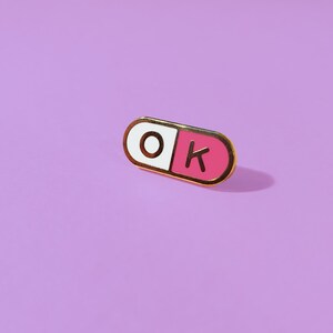 One of a pair of matching 0.75 inch; mini enamel pins in the shape of a pill; half pink, half white, with the letters O and K inside the halves. They are hard-enamel (a smooth surface, not recessed enamel), and have gold plating on the metal.