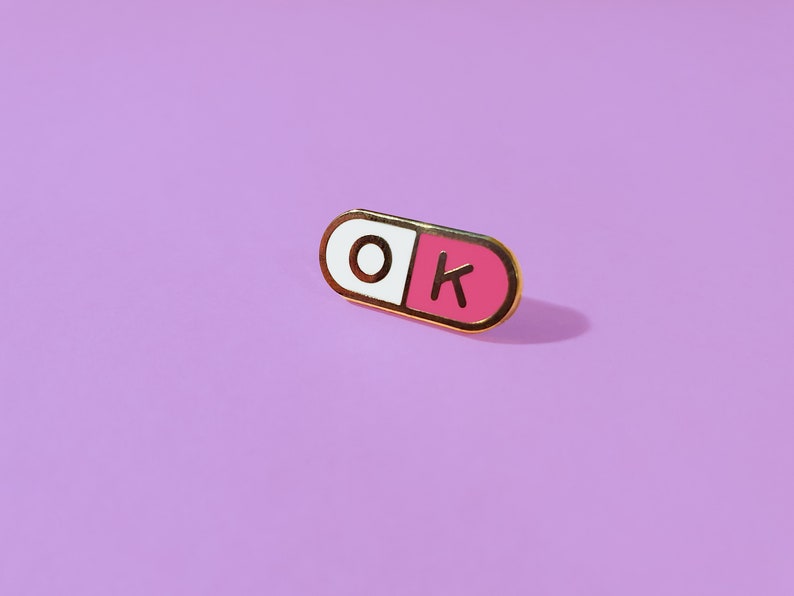 One of a pair of matching 0.75 inch; mini enamel pins in the shape of a pill; half pink, half white, with the letters O and K inside the halves. They are hard-enamel (a smooth surface, not recessed enamel), and have gold plating on the metal.