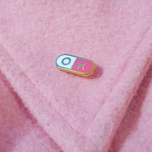 One of the enamel pins photographed on a light pink jacket coller. The pill shaped enamel pin is split in 2, with the wordK OK in the halves; O in the white half and K in the pink half.